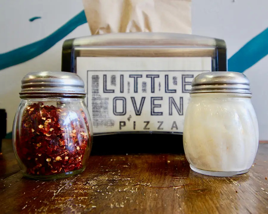 Chile flakes and parmesan shakers on a worn wooden table at Little Oven Pizza